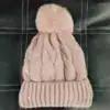 Bonnet cache oreille rose pâle à pompon, tricoté avec motif torsadé et bord côtelé.